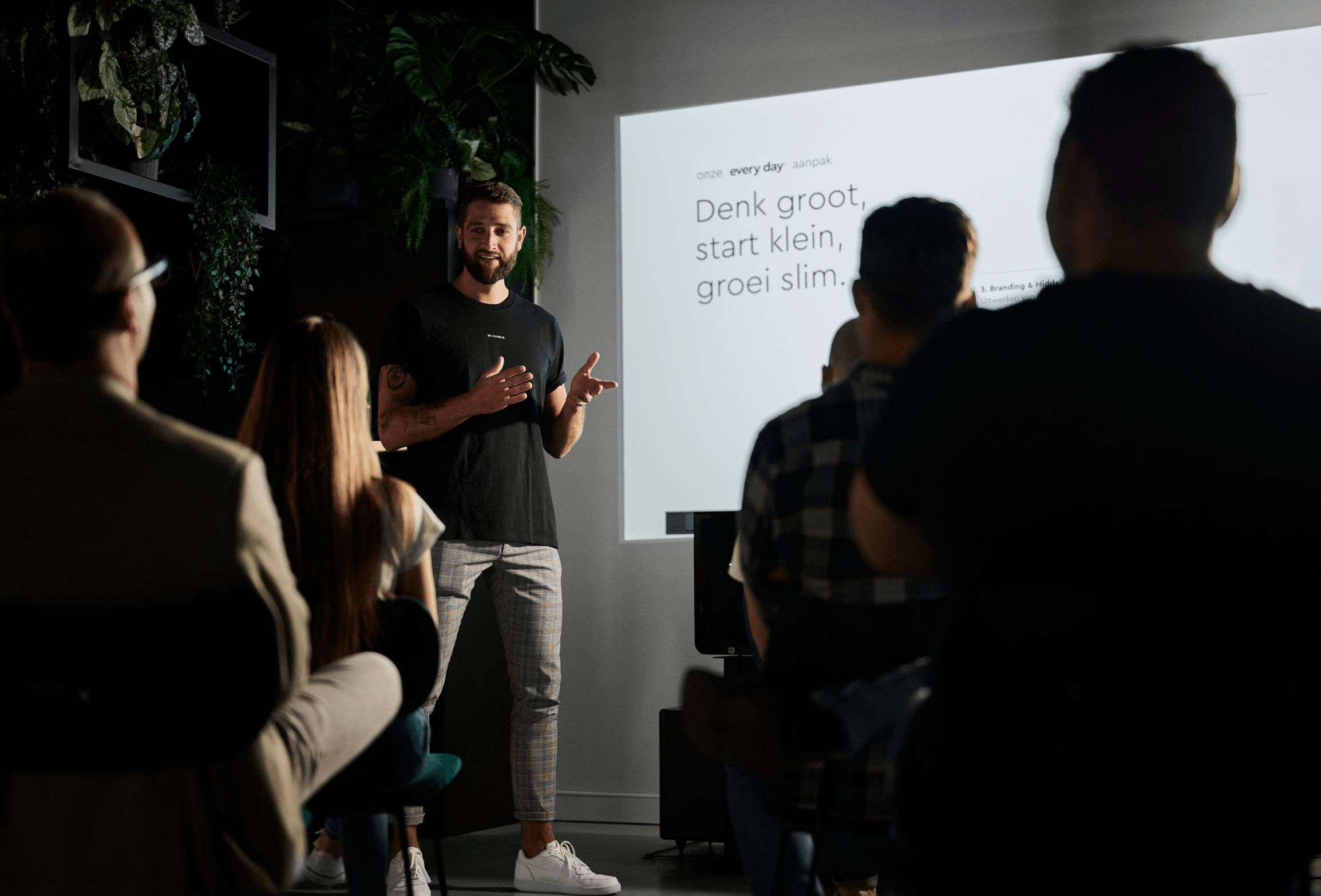 Man geeft een presentatie aan een zittend publiek met een dia waarop staat: "Denk groot, begin klein, groei slank.