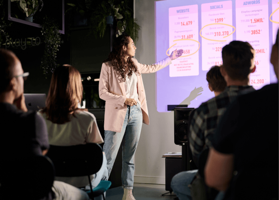 Vrouw in blazer presenteert aan publiek en wijst naar een scherm vol gegevens en grafieken.